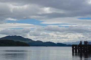 Fototapeta na wymiar Luss, Scotland: People taking selfies on a dock on Loch Lomond, with mountains on the far side of the lake. Loch Lomond is part of the Loch Lomond and The Trossachs National Park, established in 2002.