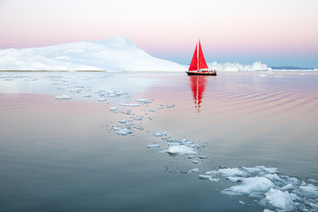 Little red sailboat cruising among floating icebergs in Disko Bay glacier during midnight sun...