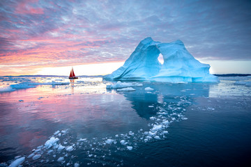 Little red sailboat cruising among floating icebergs in Disko Bay glacier during midnight sun...