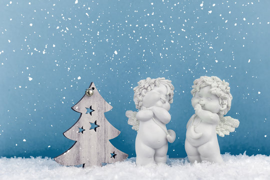 Two Christmas Baby Angels Statuettes On Snow With Wooden Christmas Tree And Copy Space