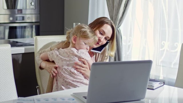 PAN of busy young mother working from home: she holding cute toddler girl and talking on phone while typing on laptop