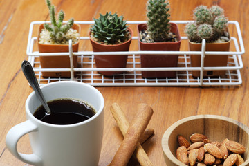 Breakfast hot black coffee in white cup, cinnamon, almond nut in wooden bowl and cactus pot plant on wood table
