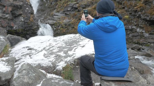 A man in sports clothes, resting near the waterfall. He takes pictures of a beautiful landscape in the mountains, on a smartphone. He leads an active and interesting life