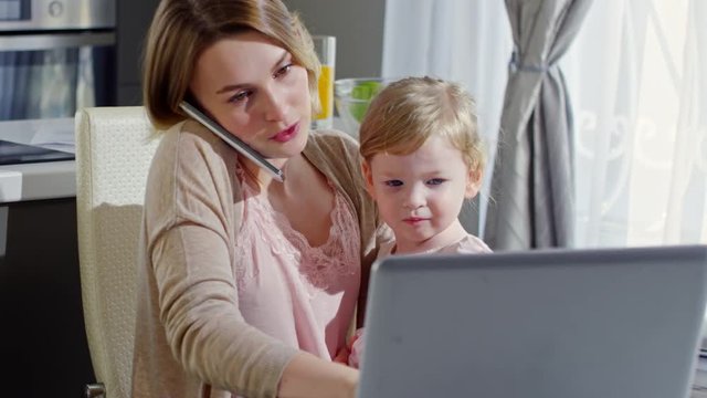 PAN of busy working mother holding cute toddler girl and talking on mobile phone while working on laptop at home