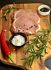 Raw fresh uncooked sliced pork fillet dish with rosemary, pepper, salt, red chili pepper, on wooden board and black background. Top side view, vertical