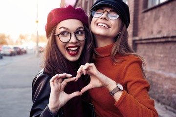 Outdoors fashion portrait young pretty best girls friends in friendly hug. Walking at the city. Posing at the street. Wearing stylish outerwear and hats. Bright make up. Positive emotions. - obrazy, fototapety, plakaty