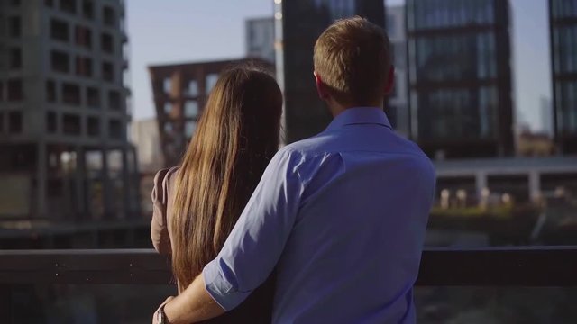 thoughtful man and woman are hugging and watching on city buildings, standing on a bridge in sunny day