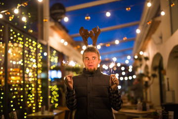 People, holidays and christmas concept - surprised man in deer's horns holding two bengals light or sparklers outdoors