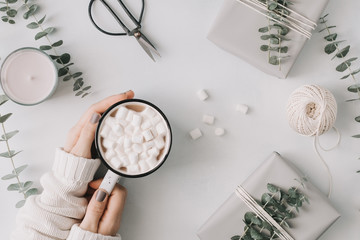 Girl's hands hold a mug with hot chocolate and marshmallow among gift boxes and eucalyptus on a white table. The concept of wintertime and wrapping gifts. Flat lay, top view, minimal style.
