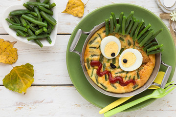 Fun food for kids - cute funny monster face made of an omelette with green beans and decorated with a boiled egg and ketchup in a pan. Healthy eating