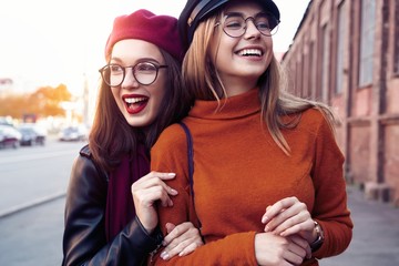 Outdoors fashion portrait young pretty best girls friends in friendly hug. Walking at the city. Posing at the street. Wearing stylish outerwear and hats. Bright make up. Positive emotions.