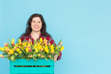 Young female florist with big box of yellow tulips over blue background with copy space