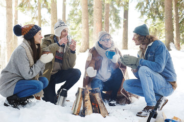 Full length portrait of four young people camping in winter forest sitting in circle round fire and...