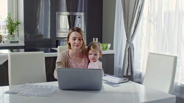 PAN of cheerful young mother holding cute toddler girl on her laps and working on laptop in kitchen