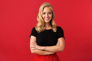 Obraz premium Portrait of beautiful blond woman 20s smiling at camera while standing with arms crossed, isolated over red background in studio