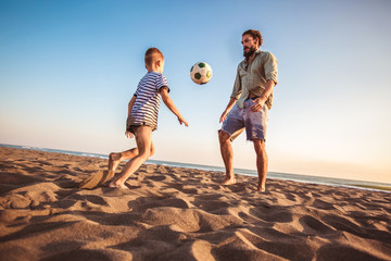 Happy father and son play soccer or football on the beach having great family time on summer holidays.
