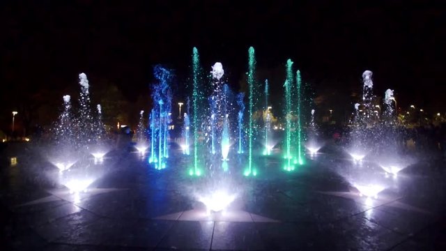 Colorful dancing fountain at night. Slow-motion footage of jets of water on a dark background. Unrecognizable people silhouettes.