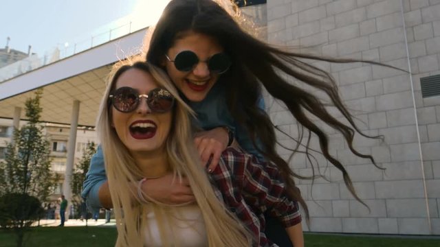 Two happy young female friends giving piggyback, laughing and walking on sunny city background. Happy young girlfriends embracing