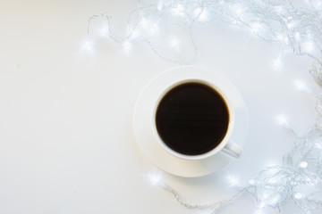 Cup of black coffee on winter white table with garland. Xmas holiday time. Top view. Copy space.