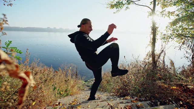 Handsome young guy standing in fighting stance and performing exercises while standing on steps near lake on sunny autumn day.