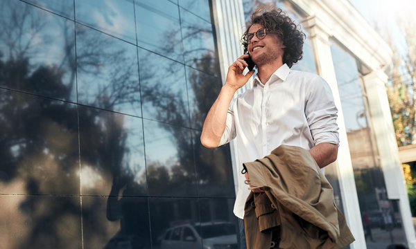 Image of handsome businessman leaning on a building's wall while standing outdoors and using smartphone. Man in suit wearing round spectacles talking with clients at his mobile phone.