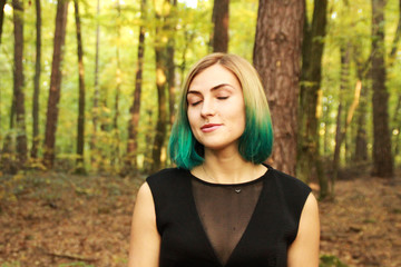 Naklejka premium The blonde girl with blue ends in a black dress stands in the woods and reads a book. The sun is warm and the sun is in the air. A portrait in the woods. Natural background. The woman is a witch