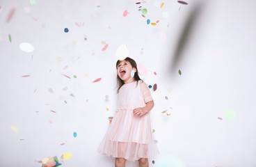 Happy beautiful little girl wearing pink dress with princess crown enjoying colorful confetti surprise falling down, posing on white studio wall. Pretty girl celebrating her birthday party, having fun