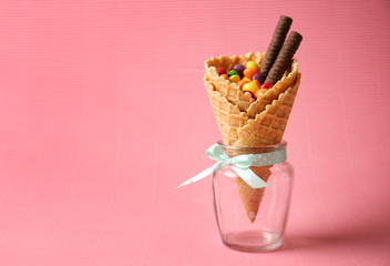 Waffle cone with delicious candies and chocolate sticks in jar on color background