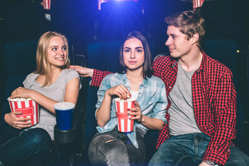 Fototapeta premium Delightful white-haired girl is looking at handsome guy. He is looking at her and keeping his hand on her shoulder. Brunette girl is looking straight forward and eating popcorn.