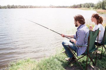 A picture of adult man and woman sitting at the river shore in this nice and beautiful sunny day. Guy is trying to catch some fish. Both of them are looking to the water.