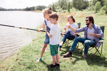 Girl and boy are holding long fish-rod together. They are concentrated on catchinf some fish in the river. Their parents are sitting behind them and watching over them.