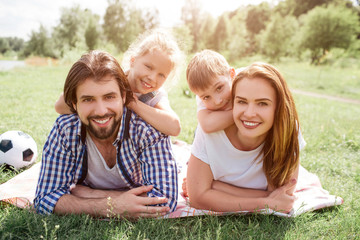 Portrait of family lying on blanket in the middle of meadow. They are looking on camera and smiling. There is a boy lying on woman's back while girl is doing the same thing on man's back.