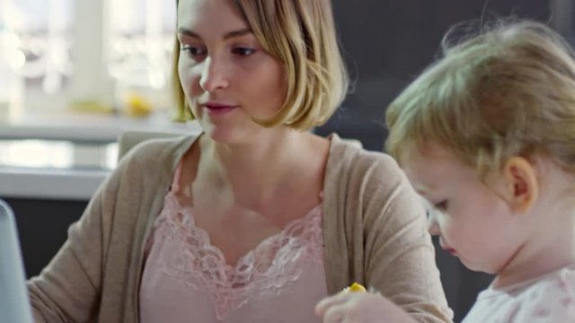 Tilt up of young mother working on laptop in kitchen and giving croissant to cute toddler girl sitting beside her and drawing with colored pencils