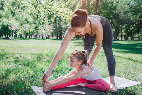 A picture of woman standing upon her daughter and teaching her to stretch. Small girl is reacing her foot with her hands and keeping other foot close to left leg. Yoga and Pilates Concept.