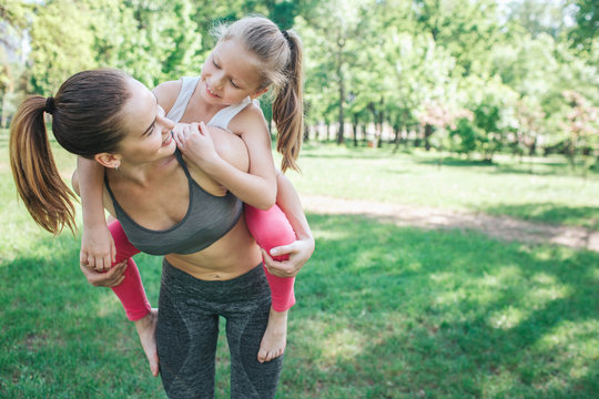 Young woman is standing outside in park and holding her daughter on her back. She is holding her on her legs and looking at smill girl. Baby is looking at mom and smiling. Yoga and Pilates Concept.