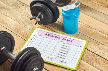 Healthy lifestyle concept. Mock up on workout and fitness dieting diary. Exercise diary sheet, blue shaker and dumbbells on a rustic wooden background