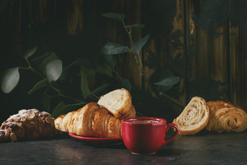 Fresh baked whole and sliced croissant with red cup of coffee espresso on black table with eucalyptus branch. Dark rustic style
