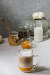 Glass of pumpkin layered spice latte with pumpkin puree, milk foam and cinnamon standing with ingredients in jars and decorative white pumpkins on white marble table with grey wall at background.