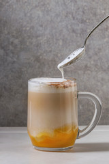 Glass of pumpkin layered spice latte with pumpkin puree, milk foam flowing from spoon and cinnamon standing on white marble table with grey wall at background. Copy space