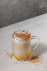 Glass of pumpkin layered spice latte with pumpkin puree, milk foam and cinnamon standing on white marble table with grey wall at background. Copy space