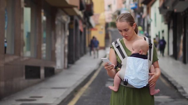Mom is standing on the street with a baby in a sling and looks at the mobile phone screen and looks through the photos