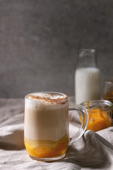 Glass of pumpkin layered spice latte with pumpkin puree, milk foam and cinnamon standing with ingredients in jars and decorative white pumpkins on crumpled table cloth with grey wall at background.