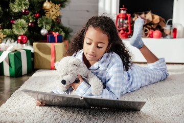 adorable african american child in pajamas with teddy bear reading book on floor at home, christmas concept