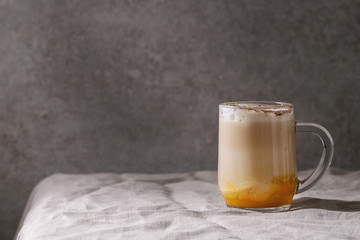 Glass of pumpkin layered spice latte with pumpkin puree, milk foam and cinnamon standing on crumpled table cloth with grey wall at background. Copy space