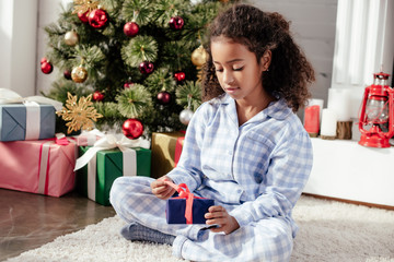 Obraz na płótnie Canvas adorable african american child in pajamas opening christmas present near christmas tree at home
