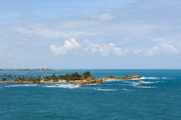 View of a tropical island in the blue sea, surrounded by reefs. Blue sky with clouds. Palm trees and beach. Waves on stone reefs.