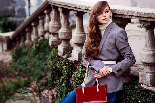 Outdoor fashion portrait of young beautiful fashionable woman wearing white turtleneck, grey checked blazer, wrist watch, holding red bag, posing in street. Copy, empty space for text
