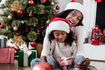 happy african american woman and daughter with gift in santa claus hats at home, newy year and christmas concept