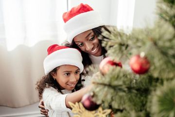 portrait of happy african american family in santa claus hats decorating christmas tree together at home