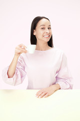 Beautiful smiling woman sitting at pink studio and looking happy holding the cup of coffee in hand. Closeup toned portrait in minimalism style